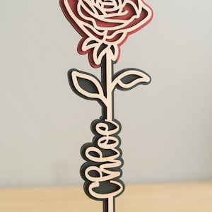 May include: A wooden rose with a red center and black outline, with the word "bless" written on the stem. The rose is standing on a heart-shaped base.