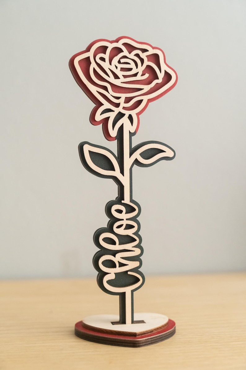 May include: A wooden rose with a red center and black outline, with the word "bless" written on the stem. The rose is standing on a heart-shaped base.