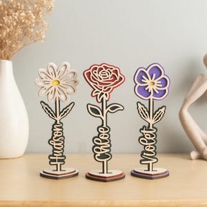 May include: Three wooden flower-shaped decorations with personalized names. The flowers are a white daisy, a red rose, and a purple pansy. Each flower has a wooden stem and base with a name written on it: Hudson, Chloe, and Violet.