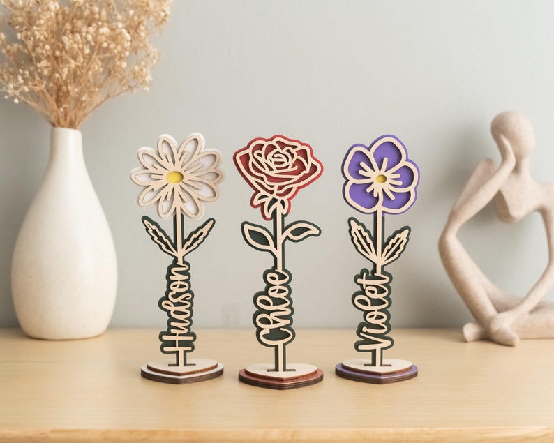 May include: Three wooden flower-shaped decorations with personalized names. The flowers are a white daisy, a red rose, and a purple pansy. Each flower has a wooden stem and base with a name written on it: Hudson, Chloe, and Violet.