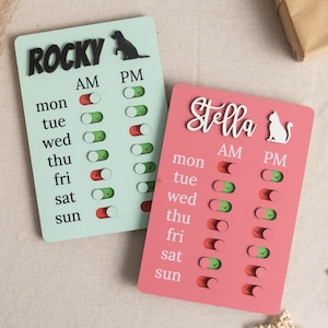 May include: Two rectangular pet schedule boards. One is light green with "Rocky" in black letters and a dog silhouette. The other is pink with "Stella" in white script and a cat silhouette. Each board has AM/PM toggles for each day of the week.