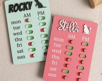 May include: Two rectangular pet schedule boards. One is light green with "Rocky" in black letters and a dog silhouette. The other is pink with "Stella" in white script and a cat silhouette. Each board has AM/PM toggles for each day of the week.