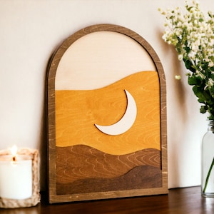 Pu&ograve; includere: Opera d'arte da parete in legno con un design paesaggistico a strati e una luna crescente. L'opera d'arte presenta una calda tavolozza di colori nei toni del beige, giallo e marrone, incorniciata da un arco in legno scuro. Una candela accesa e un vaso di fiori sono sullo sfondo.