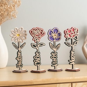 May include: Four wooden flower-shaped decorations with personalized names. The flowers are a white daisy, a red rose, a purple flower, and a pink peony. Each flower is on a wooden stand.