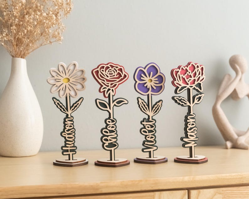 May include: Four wooden flower-shaped decorations with personalized names. The flowers are a white daisy, a red rose, a purple flower, and a pink peony. Each flower is on a wooden stand.