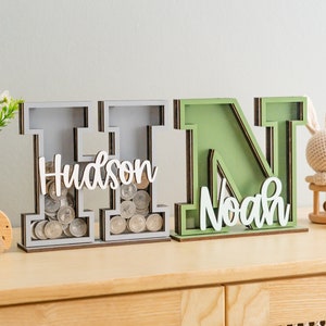 May include: Two wooden letter piggy banks, one gray and one green, with the names "Hudson" and "Noah" on them. The gray piggy bank has coins inside.