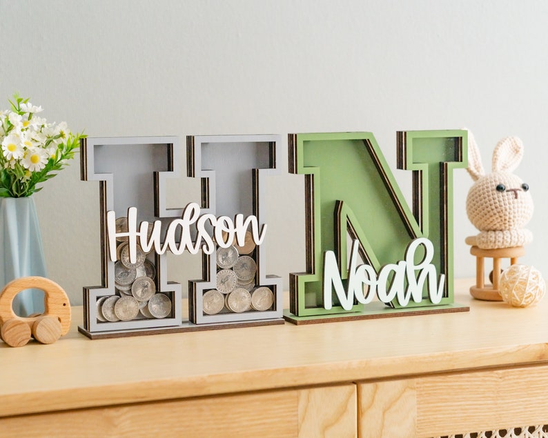 May include: Two wooden letter piggy banks, one gray and one green, with the names "Hudson" and "Noah" on them. The gray piggy bank has coins inside.