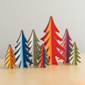 May include: A collection of eight freestanding wooden tree ornaments in various sizes and colours, including yellow, light blue, dark blue, white, green, red, orange, and purple. Each tree features a jagged, stylized silhouette with visible wood edges.
