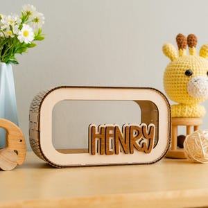 May include: A wooden box with a cutout window and the name "HENRY" spelled out in brown letters. The box is sitting on a wooden table next to a yellow crocheted giraffe toy.