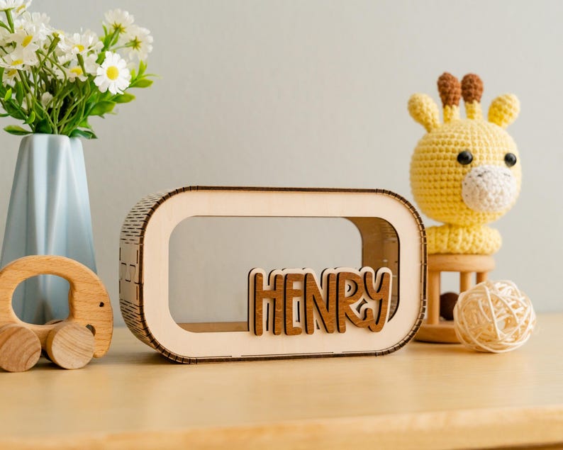 May include: A wooden box with a cutout window and the name "HENRY" spelled out in brown letters. The box is sitting on a wooden table next to a yellow crocheted giraffe toy.