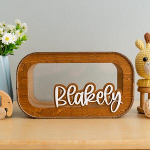 May include: A wooden box with a clear window and the name "Blakely" written in white letters. The box is sitting on a wooden surface.