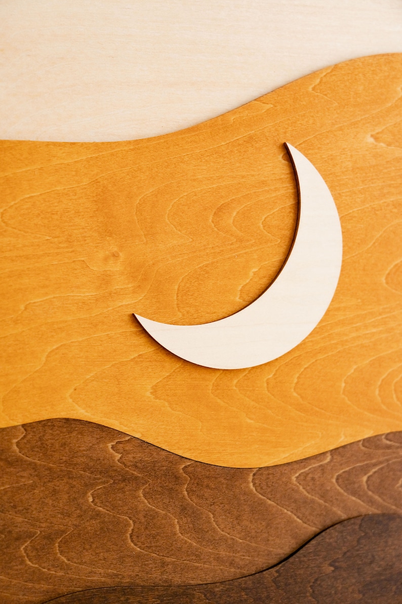 Pu&ograve; includere: Arte da parete in legno raffigurante una luna crescente su uno sfondo a strati di colori caldi. Il design include sfumature di beige chiaro, giallo dorato e marrone scuro, creando un tema tramonto o paesaggio desertico.
