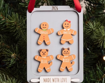 May include: A rectangular gray wooden ornament with a red ribbon hanger, featuring the year 2026 at the top and four gingerbread-style figures labeled Daniel, Amelia, Blue, and Roxie. A white plaque at the bottom reads MADE WITH LOVE.
