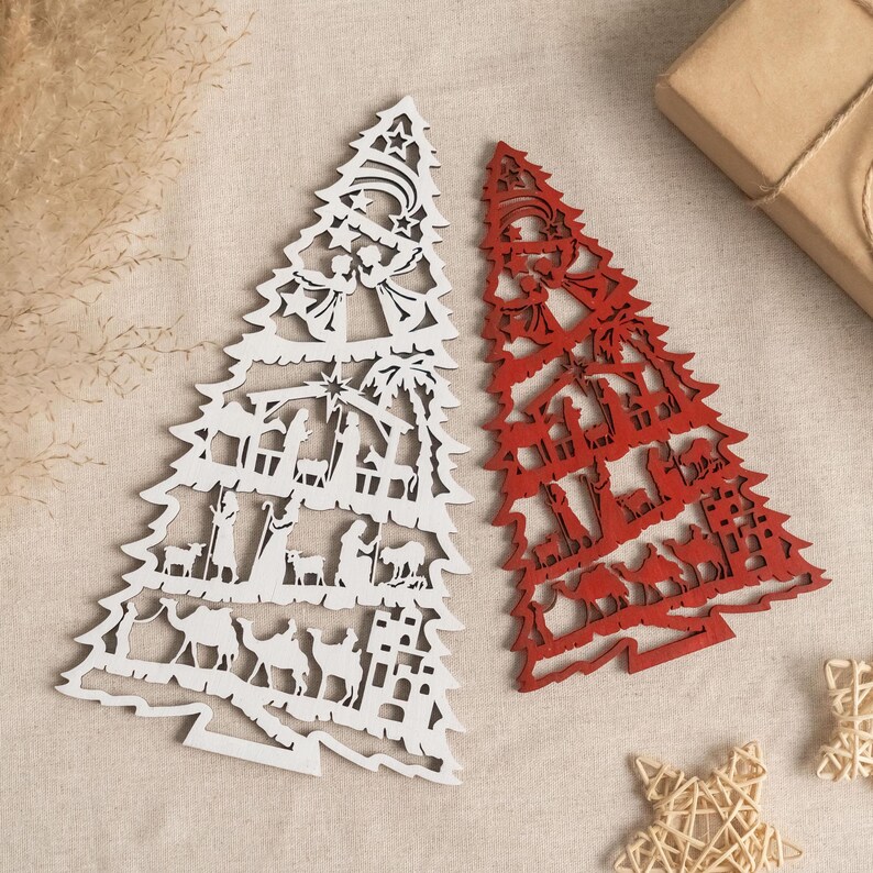 May include: Two wooden Christmas tree ornaments, one white and one red, with a nativity scene design. The ornaments are cut out and have a detailed design.