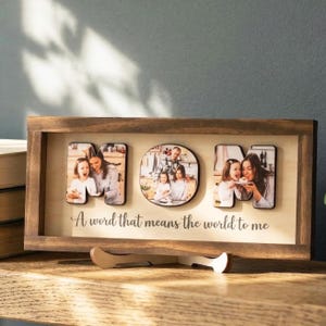 Puede incluir: Un expositor de fotos enmarcado en madera con la palabra "MOM" escrita con fotos individuales. La pantalla incluye la frase "A word that means the world to me". El marco es marrón y se encuentra sobre una superficie de madera.