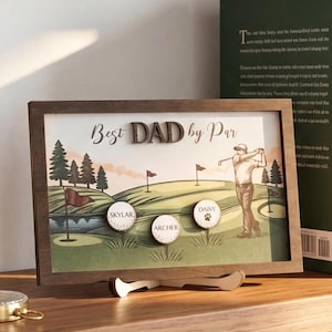 May include: A wooden framed golf-themed artwork with the text "Best DAD by Par." The artwork features a golf course scene with a golfer, flags, and personalized golf ball markers with names. The frame has a stand.