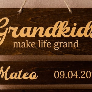 May include: A dark brown wooden hanging sign with light-colored lettering. The top board reads Grandkids make life grand. A lower board is attached with metal rings, featuring the name Mateo and the date 09.04.2014 in a light-colored font.