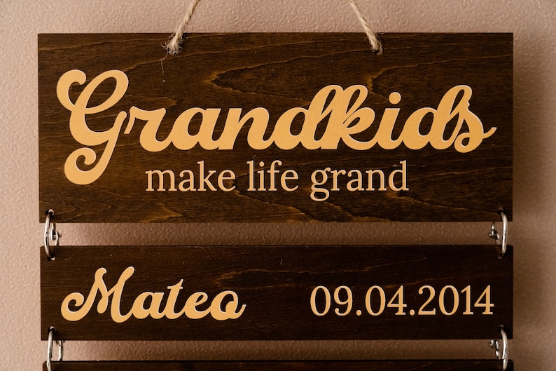 May include: A dark brown wooden hanging sign with light-colored lettering. The top board reads Grandkids make life grand. A lower board is attached with metal rings, featuring the name Mateo and the date 09.04.2014 in a light-colored font.