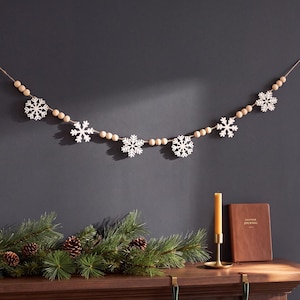 May include: A decorative garland featuring five white wooden snowflake cutouts interspersed with natural light brown wooden beads strung on a thin cord. The snowflake shapes are intricate and symmetrical, creating a repeating pattern along the string.