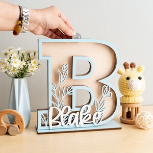 May include: Light blue wooden letter B piggy bank with a cutout floral design and the name "Blake" written in white. The piggy bank is on a wooden surface with a small stuffed giraffe and a wooden toy.