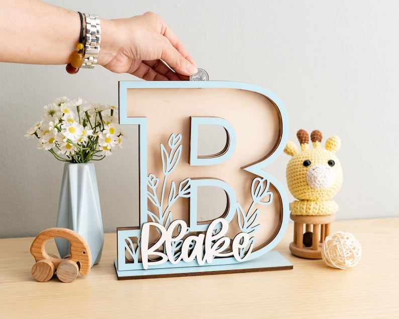 May include: Light blue wooden letter B piggy bank with a cutout floral design and the name "Blake" written in white. The piggy bank is on a wooden surface with a small stuffed giraffe and a wooden toy.