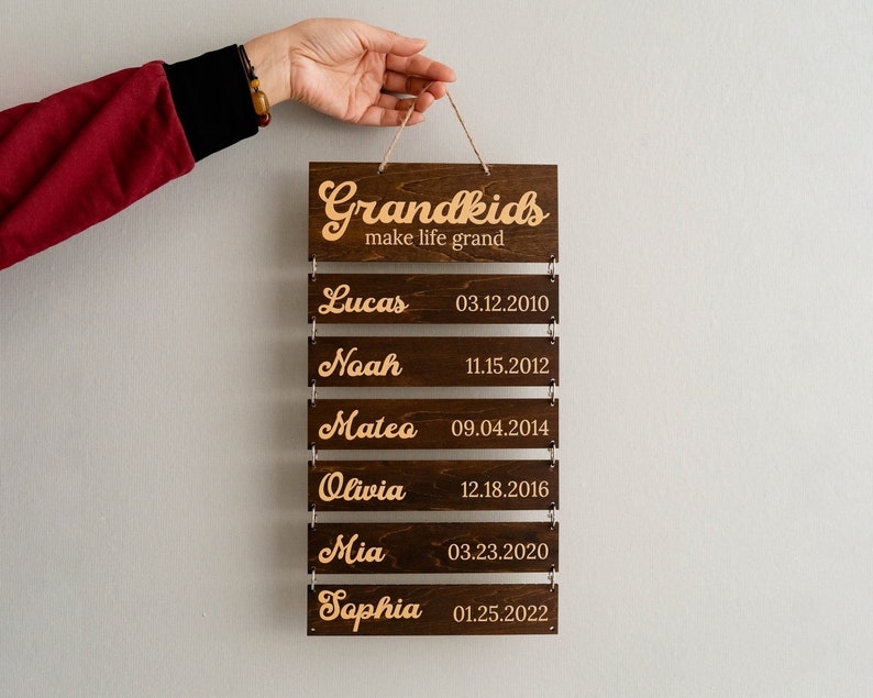 May include: A dark brown wooden wall hanging sign with six horizontal slats connected by metal rings. The top slat reads Grandkids make life grand. The five lower slats feature engraved names and dates: Lucas 03.12.2010, Noah 11.15.2012, Mateo 09.04.2014, Olivia 12.18.2016, Mia 03.23.2020, and Sophia 01.25.2022.