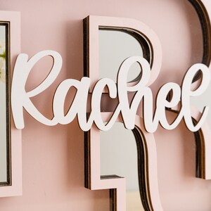 May include: A pink wooden letter "R" with a mirror inside. The letter is decorated with a white cursive name "Rachel".