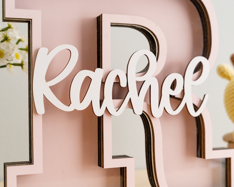May include: A pink wooden letter "R" with a mirror inside. The letter is decorated with a white cursive name "Rachel".