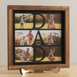 Puede incluir: Marco de fotos de madera marrón oscuro con la palabra "DAD" recortada, que muestra fotos de un padre y un niño. El marco tiene un fondo negro. Las fotos muestran a un padre y un niño jugando con un avión de juguete.