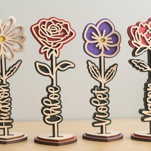 May include: Four wooden flower sculptures with different colored flowers and names written on the stems. The flowers are a white daisy, a red rose, a purple pansy, and a pink peony. The names are Hudson, Chloe, Violet, and Mason.