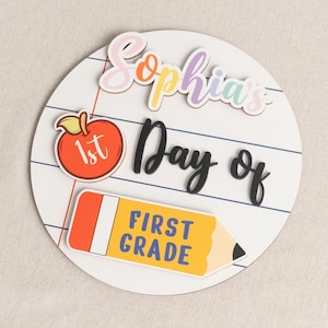 Könnte beinhalten: Ein rundes weißes Holzschild mit einem linierten Papiermuster. Der Text lautet Sophia's 1st Day of FIRST GRADE. Es enthält einen roten Apfel-Ausschnitt mit dem Text 1st und einen gelben Bleistift-Ausschnitt mit dem Text FIRST GRADE in blauen Buchstaben.