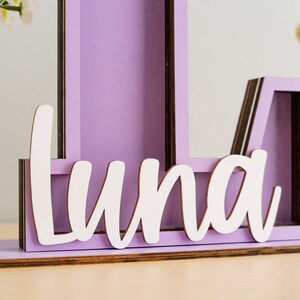 May include: A purple wooden letter L with a white cutout of the name "Luna" on the bottom. The letter is made of multiple layers of wood and has a rustic look.