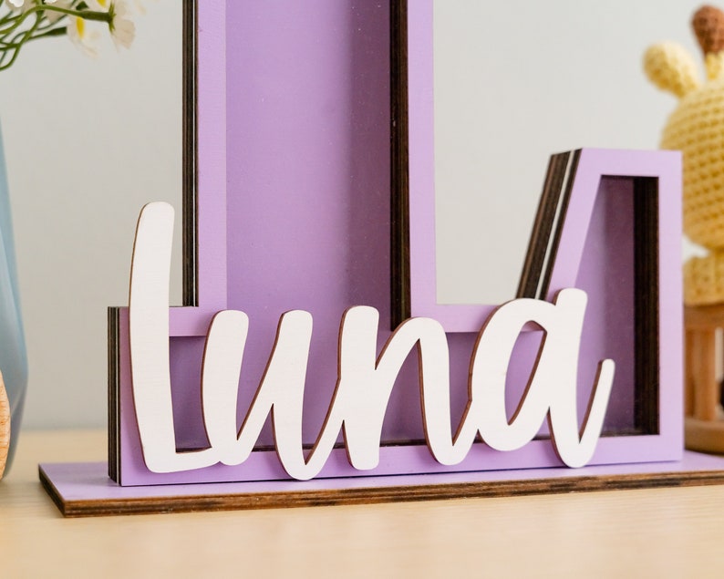 May include: A purple wooden letter L with a white cutout of the name "Luna" on the bottom. The letter is made of multiple layers of wood and has a rustic look.