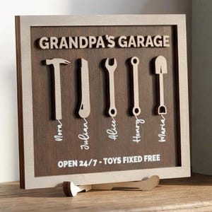 Könnte beinhalten: Holzschild mit dem Text "Grandpa's Garage" und Werkzeugausschnitten. Das Schild enthält die Namen Nora, Julian, Alice, Henry und Maria. Auf dem unteren Teil des Schildes steht "Open 24/7 - Toys Fixed Free".