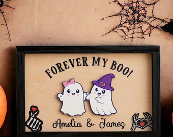 Może przedstawiać: Drewniany znak z czarną ramą i tekstem "Forever My Boo!" z dwoma kreskówkowymi duchami trzymającymi się za ręce. Duchy są białe z czarnymi oczami i ustami. Znak zawiera również imiona "Amelia & James" napisane pod duchami.