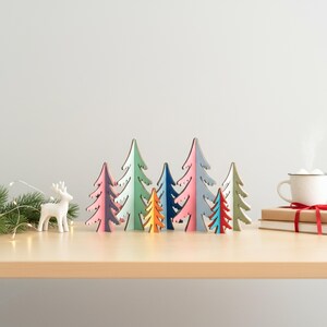 May include: A collection of seven wooden tree ornaments in various sizes and colours, including shades of pink, blue, green, orange, and red. A small white deer figurine stands to the left, and a white mug sits on a stack of books to the right.
