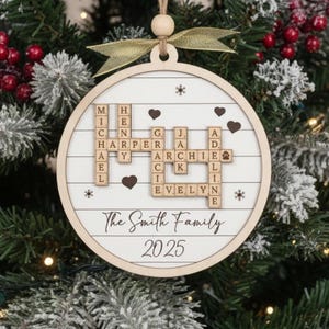 May include: A round, wooden Christmas ornament with a white center and a natural wood border. The ornament features the names of family members in a crossword puzzle design, along with the text "The Smith Family 2025". It is decorated with hearts and snowflakes.