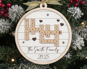 May include: A round wooden Christmas ornament featuring a white background with horizontal lines. It displays a crossword-style arrangement of names including Michael, Henry, Harper, Gracie, Jack, Archie, Adeline, and Evelyn. The text reads The Smith Family 2025, accented with small hearts and snowflakes, topped with a gold ribbon.