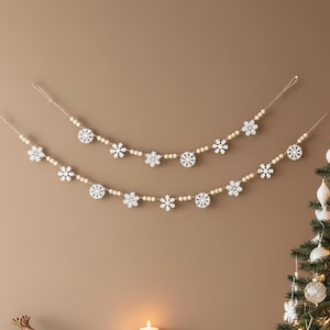 May include: Two decorative garlands featuring white wooden snowflake cutouts interspersed with natural light-colored wooden beads. The garlands are strung on thin twine and arranged in a draped, curved pattern against a solid neutral wall.