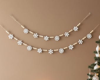 May include: Two decorative garlands featuring white wooden snowflake cutouts interspersed with natural light-colored wooden beads. The garlands are strung on thin cord and arranged in a draped, curved pattern against a solid wall.