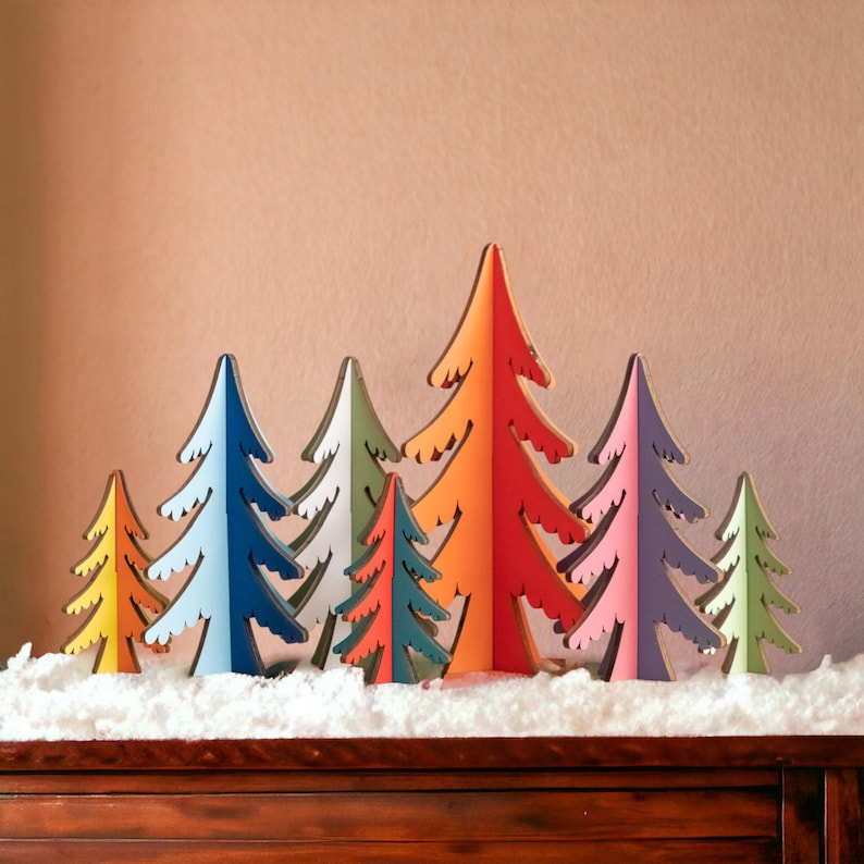 May include: A collection of eight freestanding wooden tree ornaments in various sizes and colours, including yellow, blue, white, teal, orange, red, purple, and light green, arranged in a row on a layer of white artificial snow.