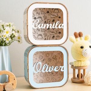 Puede incluir: Dos cajas de madera cuadradas apiladas con esquinas redondeadas. La caja superior es blanca con el nombre Camila en letra cursiva y un fondo con patrones de dinosaurios. La caja inferior es azul claro con el nombre Oliver en letra cursiva.