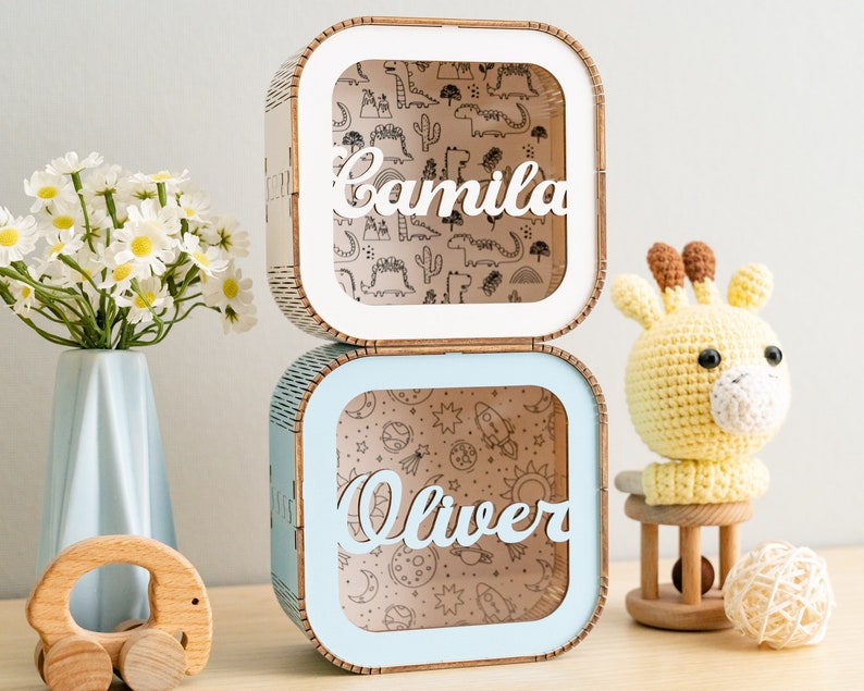 Puede incluir: Dos cajas de madera cuadradas apiladas con esquinas redondeadas. La caja superior es blanca con el nombre Camila en letra cursiva y un fondo con patrones de dinosaurios. La caja inferior es azul claro con el nombre Oliver en letra cursiva.