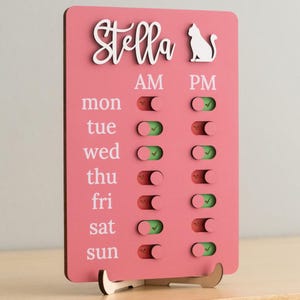 Op de afbeelding: Roze houten schema bord voor huisdierverzorging met de naam "Stella" in witte letters, een wit katten silhouet en tuimelschakelaars voor AM en PM. De dagen van de week staan vermeld met schakelaars.