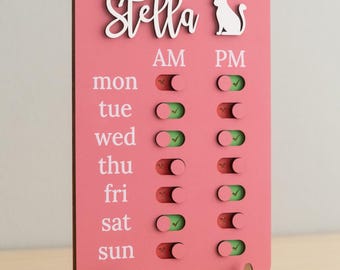 May include: Pink wooden pet care schedule board with the name "Stella" in white script, a white cat silhouette, and toggle switches for AM and PM. The days of the week are listed with toggle indicators for each time slot.