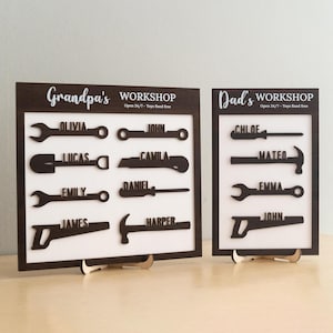 Könnte beinhalten: Zwei Werkstattschilder aus Holz, eines mit der Aufschrift "Grandpa's WORKSHOP" und das andere mit "Dad's WORKSHOP". Jedes Schild zeigt Werkzeugförmige Ausschnitte mit Namen. Die Schilder stehen auf Holzständern, mit dem Text "Open 24/7 - Toys fixed free."