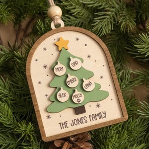 May include: An arched wooden Christmas ornament featuring a green tree with a yellow star. Small circular wooden tags are attached to the tree, engraved with the names MOM, DAD, EMERY, LANA, ALEX, and MOLLY, with a paw print. The text THE JONES FAMILY is printed below.
