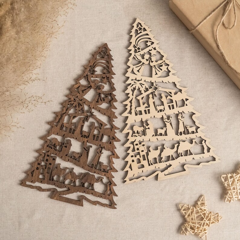 May include: Two wooden Christmas tree ornaments with a nativity scene design. The tree on the left is brown and the tree on the right is natural wood.