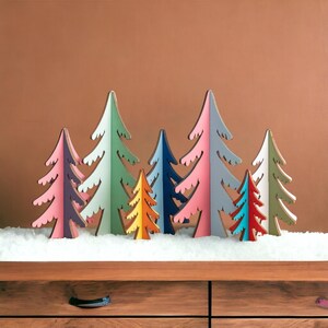 May include: A collection of eight stylised wooden tree ornaments in various colours including pink, purple, green, orange, blue, and white. The trees feature jagged, pointed branches and are arranged in a row, standing upright on a white surface.