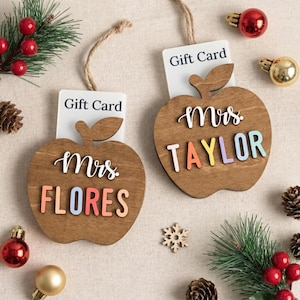 May include: Two wooden apple-shaped gift card holders with jute twine loops. Each apple features the white script text Mrs. and colorful block letters spelling FLORES or TAYLOR. The holders are designed to display a white gift card.